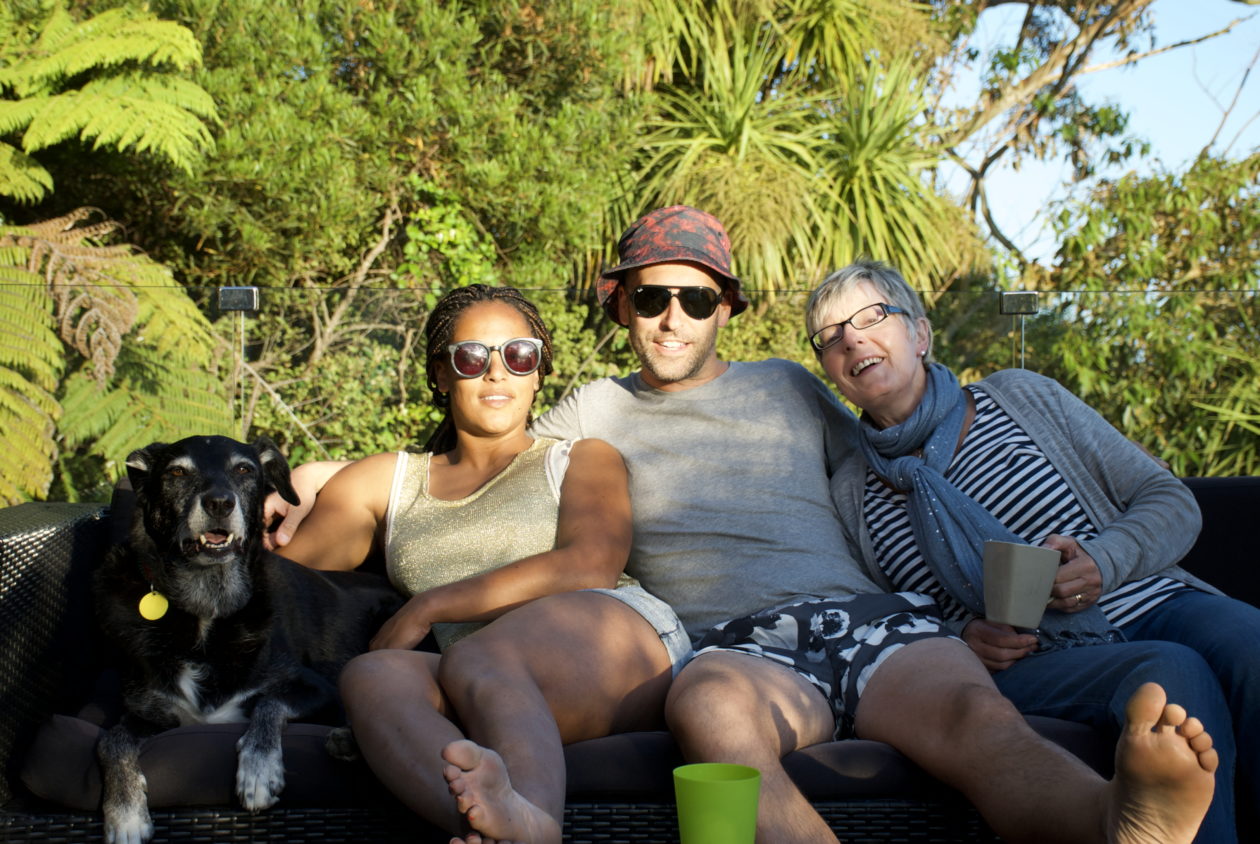 a dog, young women and young man huddling together with an older lady holding drinking mugs smiling relaxing on an outdoor couch.