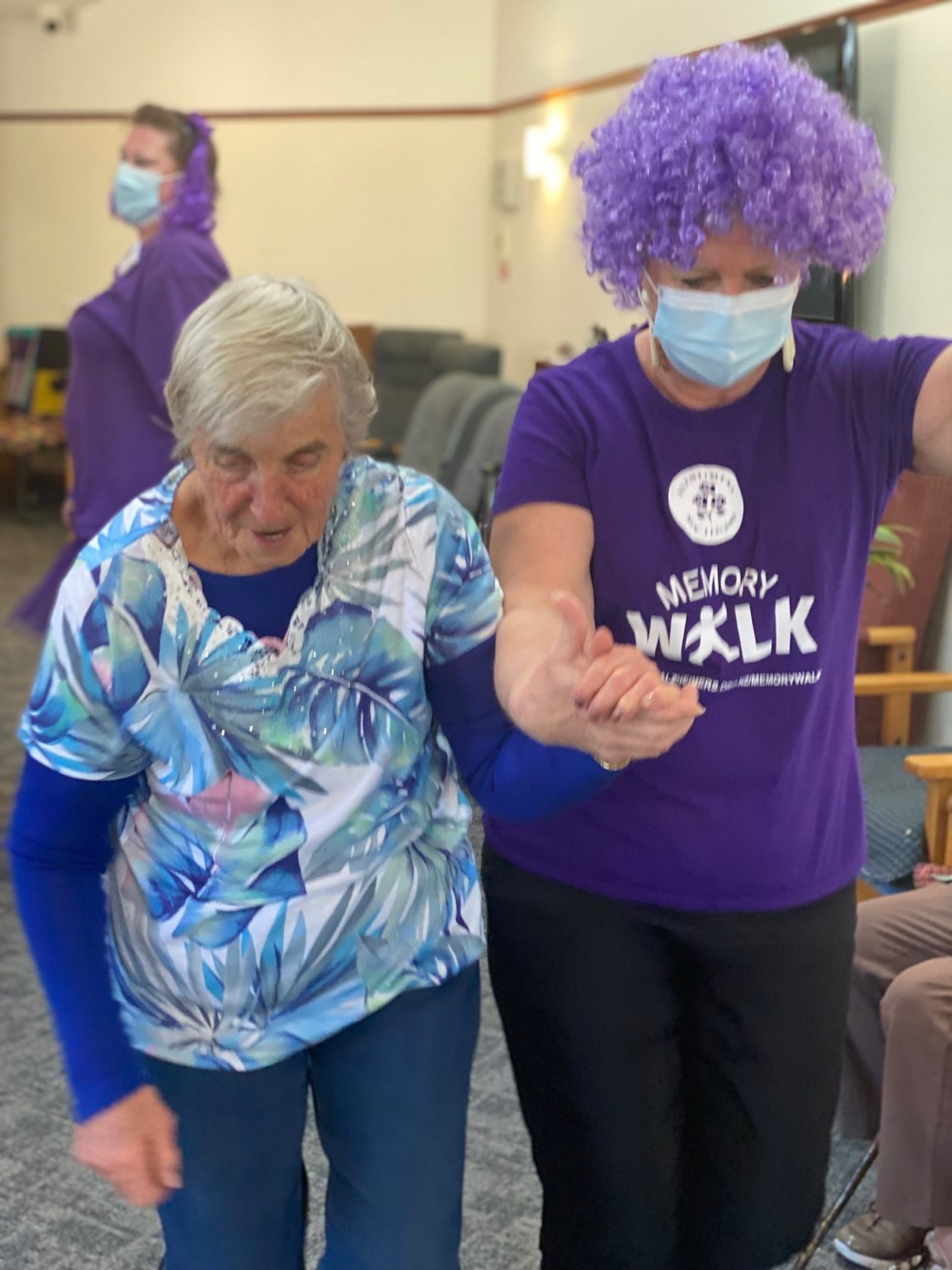 one women wearing a purple wig and a purple shirt holds an elderly women's hand leading her.