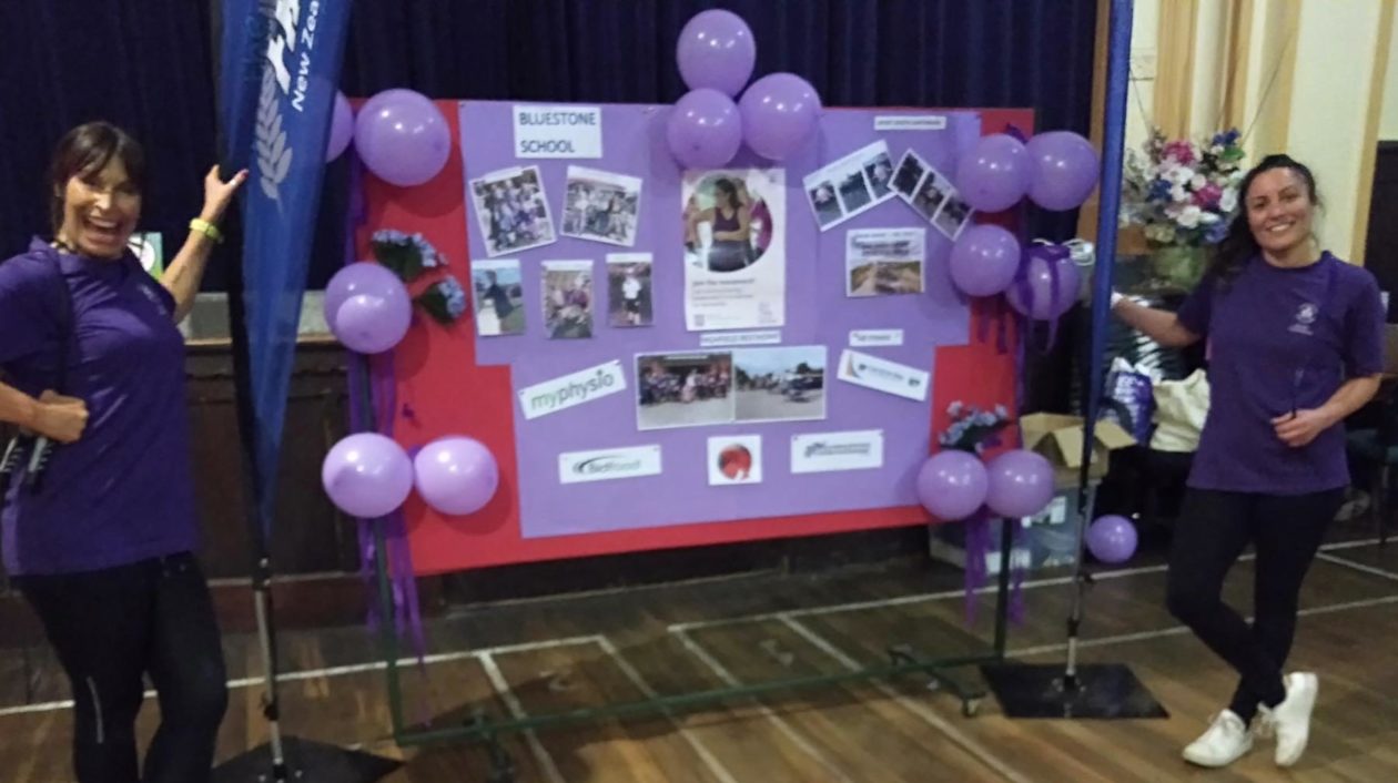 2 women in purple shirts stand posing next to a noticeboard decorated with purple decorations and posters 