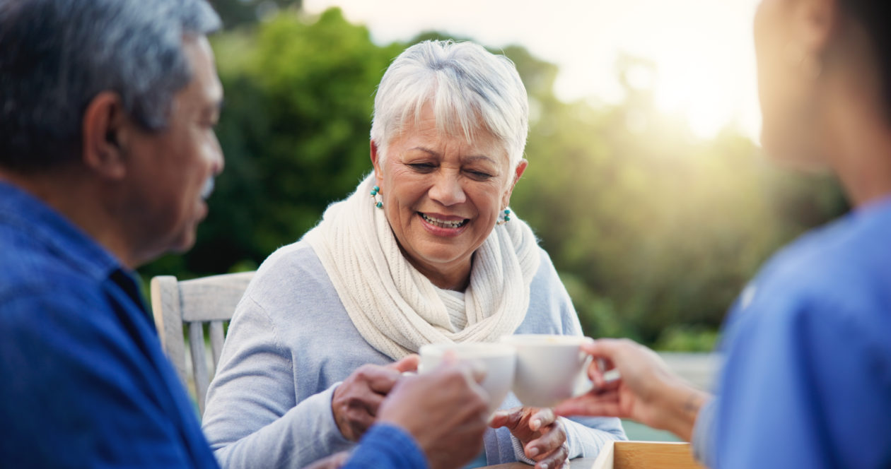 a group of aged people sitting on comfortable arm chairs laughing and holding mugs.
