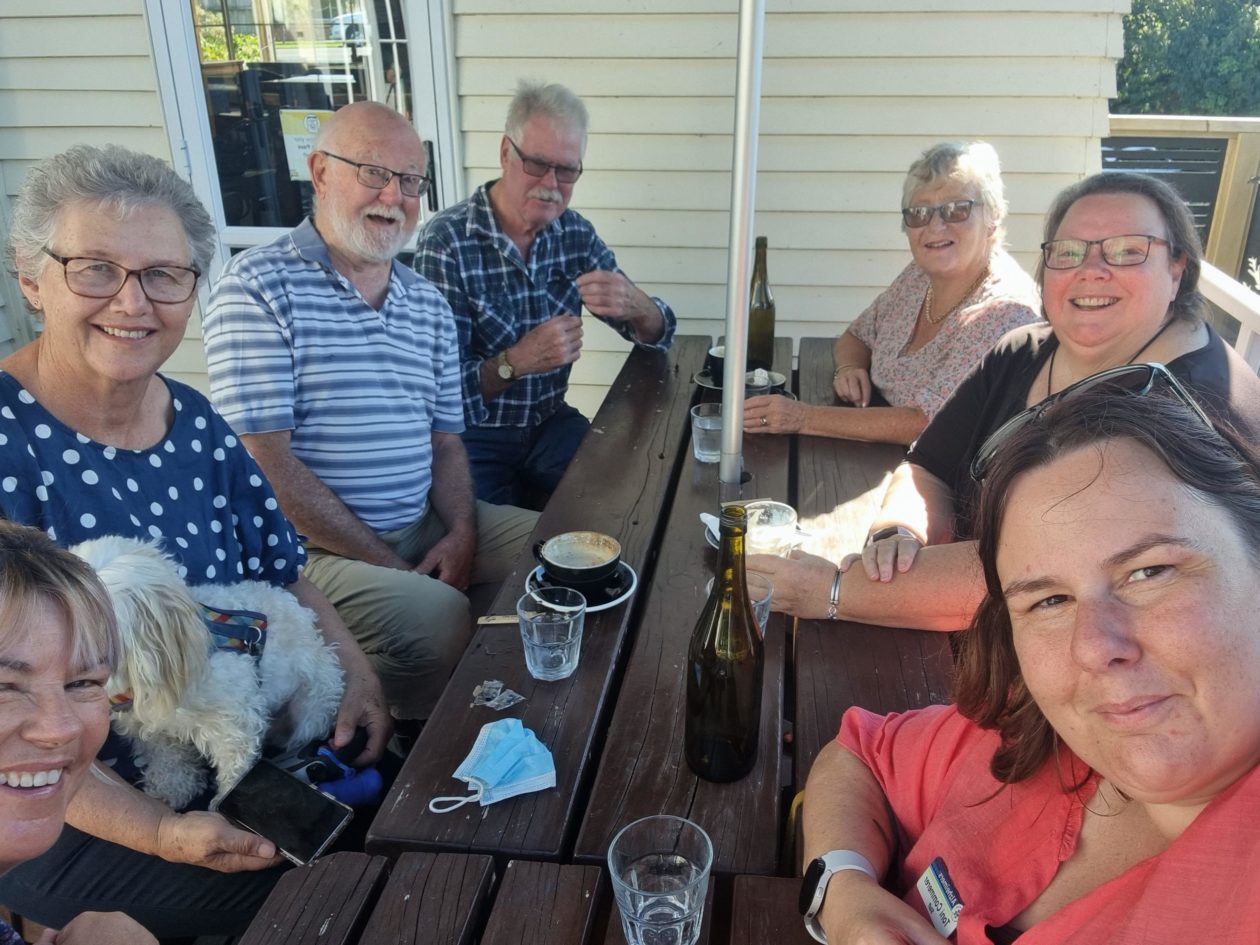 photo of several group members enjoying coffee in the sunshine on a outdoor table at the Stumble Inn, New Plymouth. All have smiles on their faces and one person has a dog on her lap.