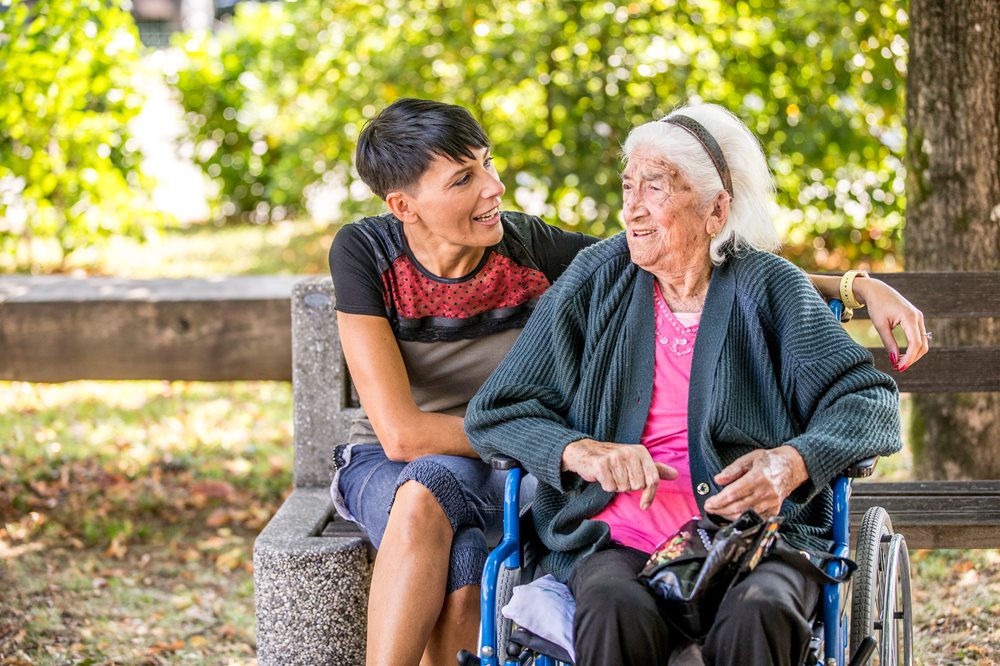 Senior woman in wheelchair with a family member who is sitting on a park bench.