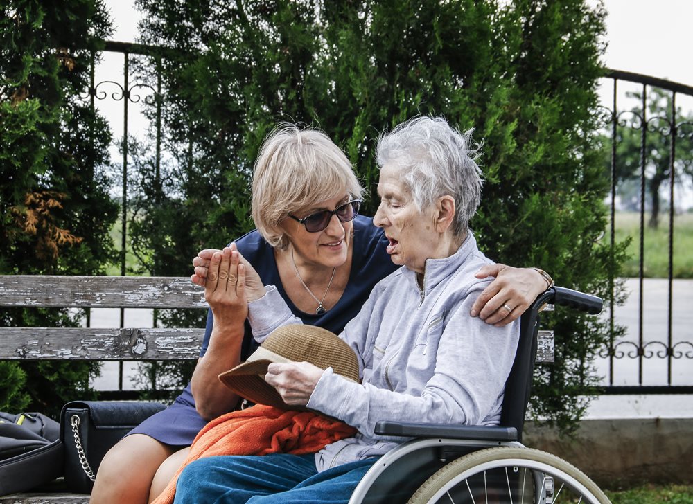 Senior woman in her wheel chair and another woman with her arm around her while holding her hand.